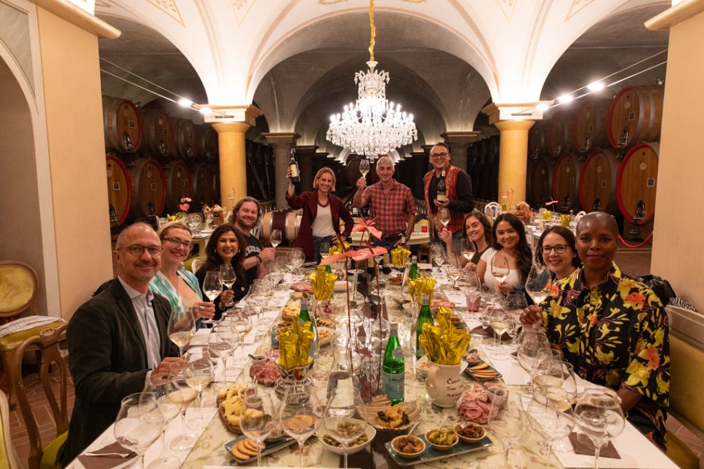 Un gruppo di giornalisti americani è seduto a un tavolo apparecchiato in una cantina, brindando durante una degustazione di vini Lugana.